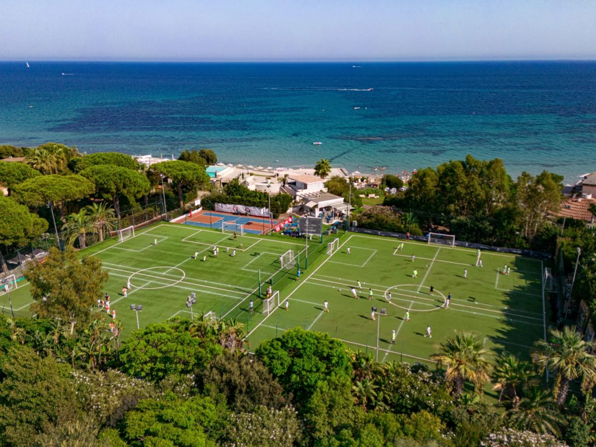 Aerial view of two green football pitches and a multi-sport court just back from a white-sand beach and turquoise sea