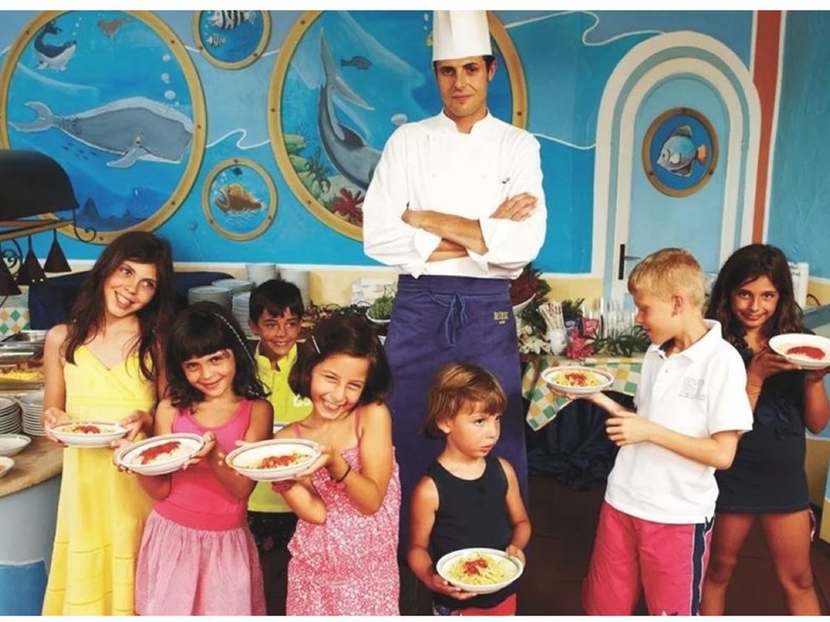 A chef behind a buffet counter serving young children holding plates, in a room with a marine mural