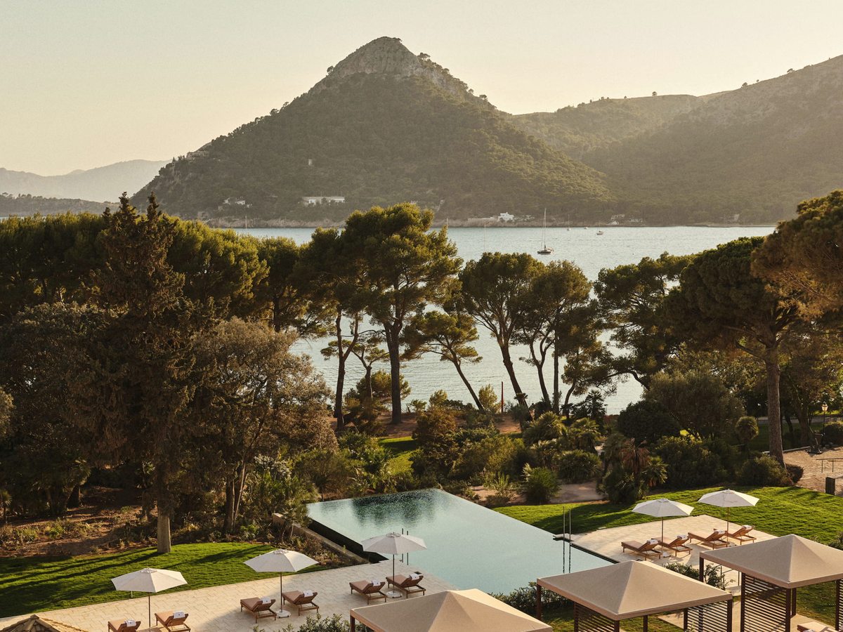 An infinity-edge pool with rows of sun loungers and cream parasols on a lawn, pine trees around it and a pyramid-shaped mountain across the bay behind.