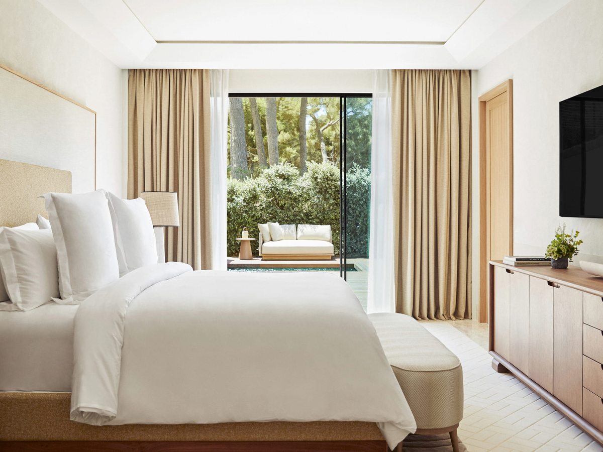 A room with a king bed and sliding glass doors opening onto a terrace with a sofa and a small plunge pool, backed by garden greenery.