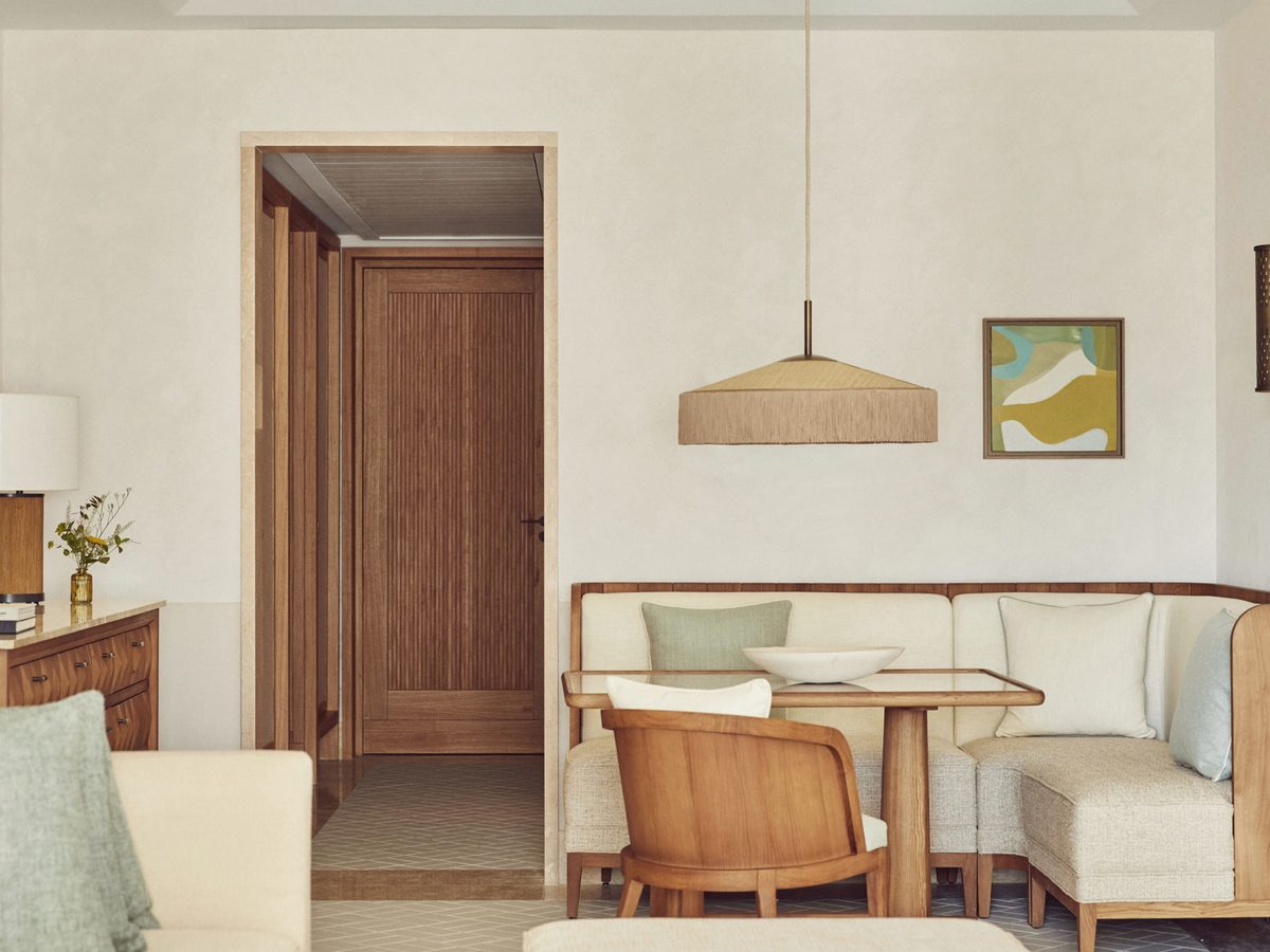 A suite living room with a built-in banquette, a glass-topped dining table and an armchair, with a doorway through to the rest of the suite.