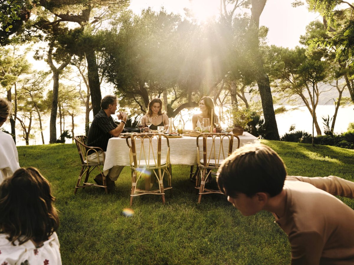 Two adults at a linen-set table on a grassy clearing among pine trees, with children on the grass and the bay behind in low sun.