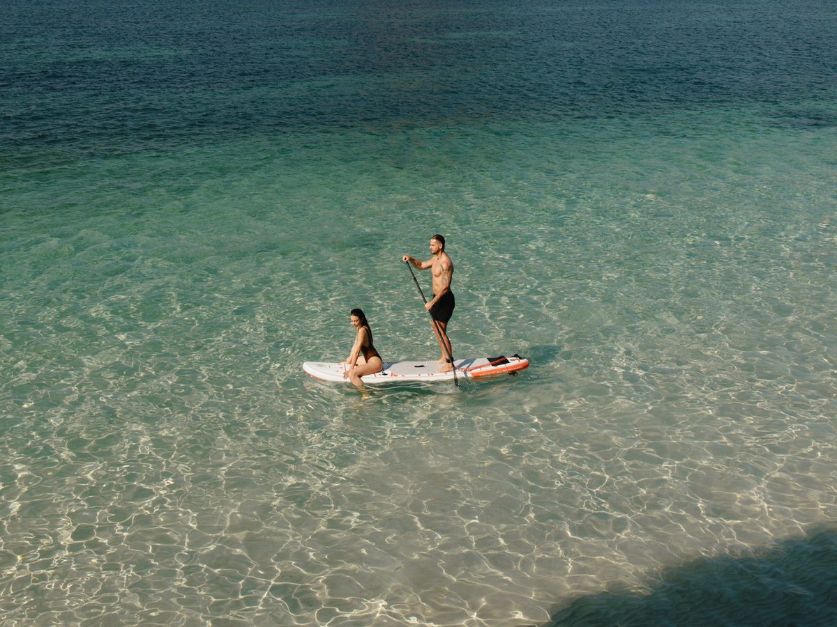 Two people on a stand-up paddleboard in clear, shallow turquoise water off the beach.