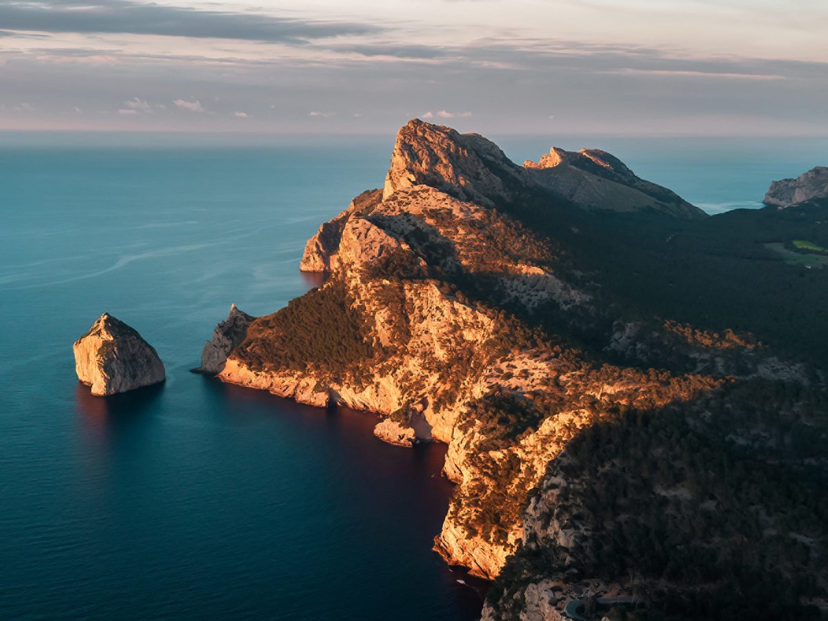 Rugged limestone cliffs and a mountainous headland dropping to the sea, with an offshore rock stack, in warm evening light.