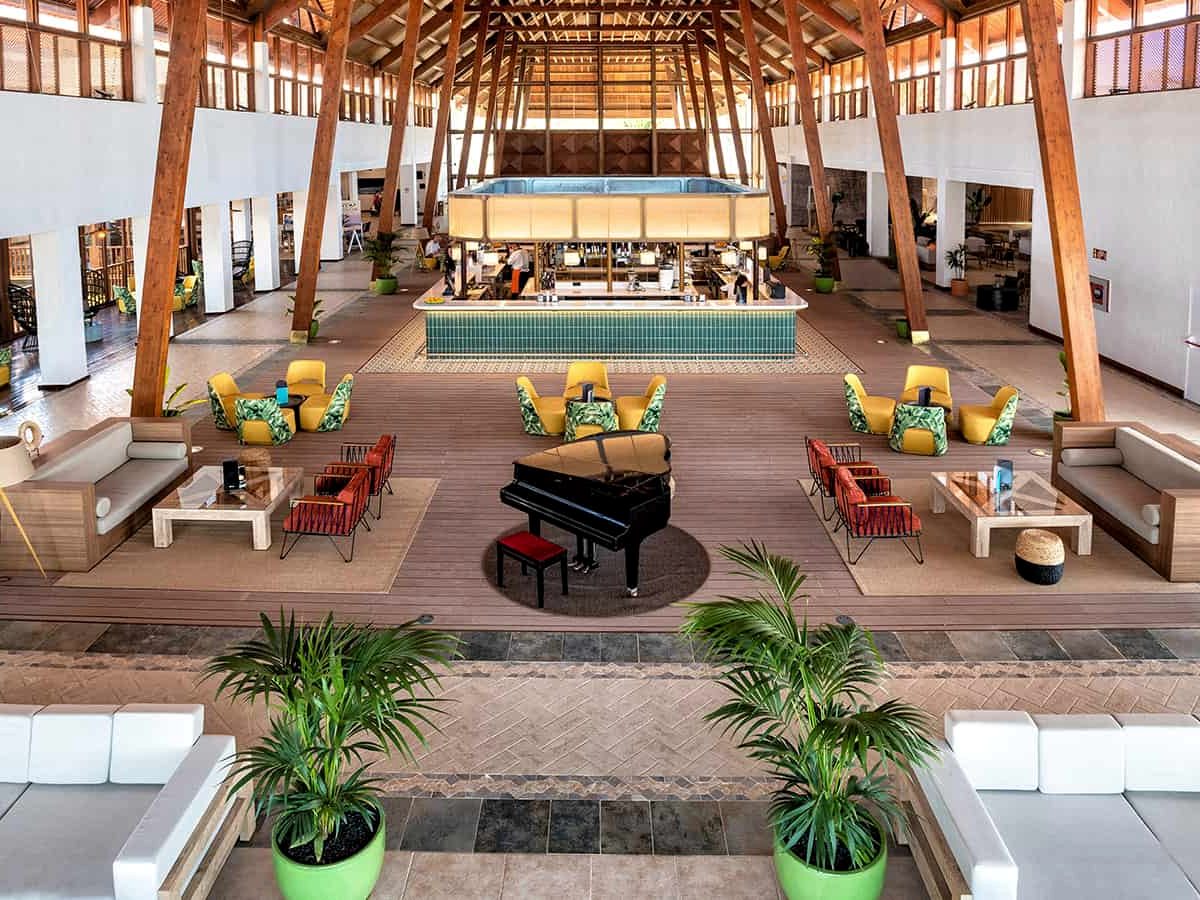 A large open-sided lobby under a high timber roof, with a black grand piano and clusters of green and yellow armchairs.