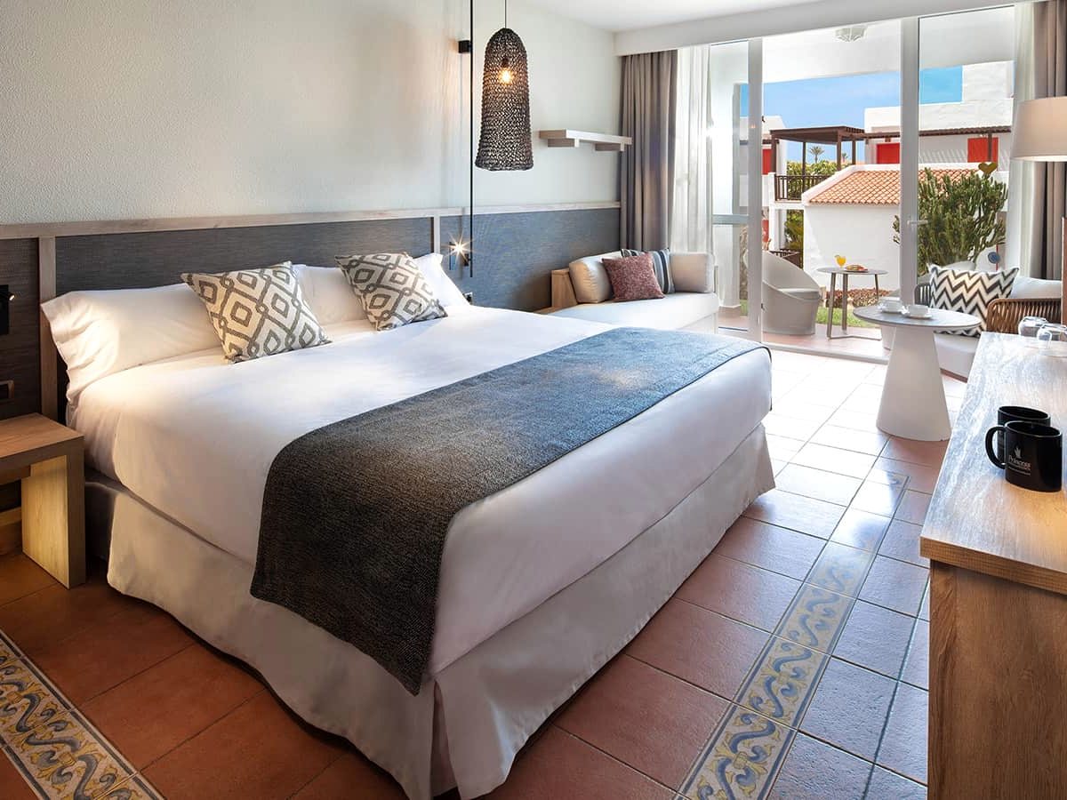 A room with a large bed dressed in white and grey, terracotta tiled floor, and a sliding door onto a terrace over the gardens.