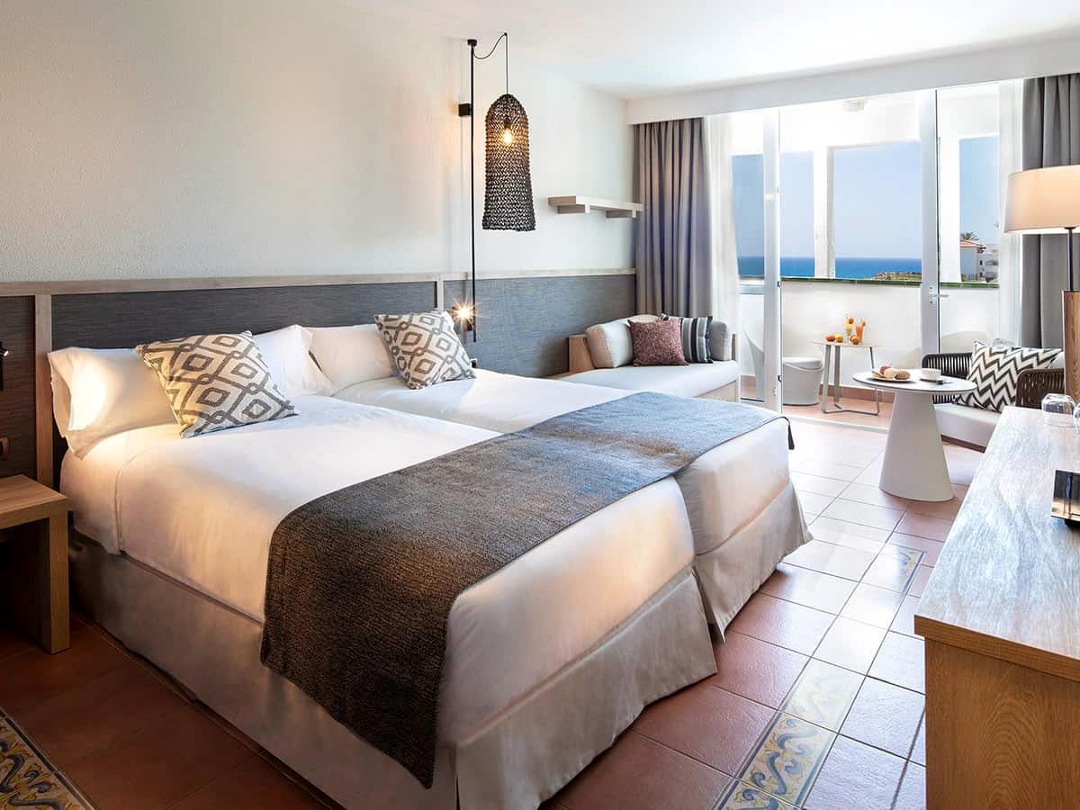 A room with a double bed and a sofa, a lamp and side table, and a balcony door with a sea view.