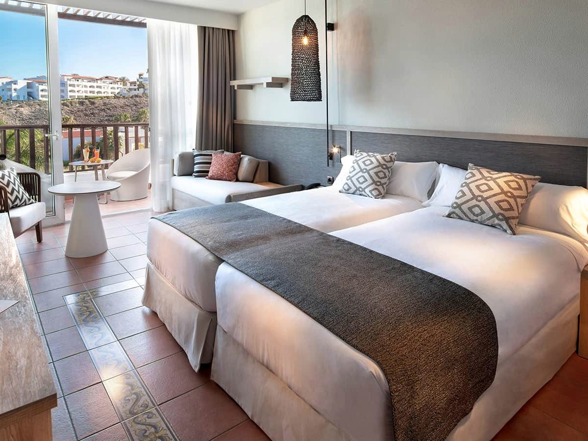 A room with two single beds pushed together, a seating corner and a balcony looking out to the sea.