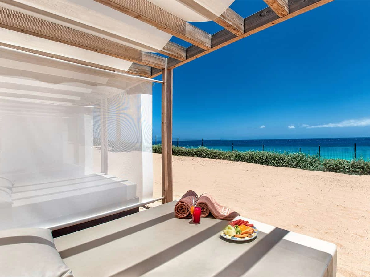 A white curtained daybed on golden sand with a fruit platter, the sea in the background.