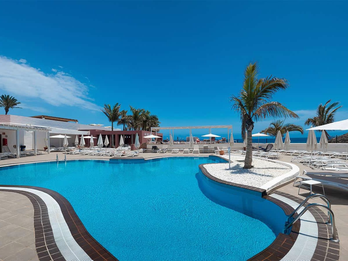 A large curved blue pool edged with sun loungers and a few palm trees under a clear sky.