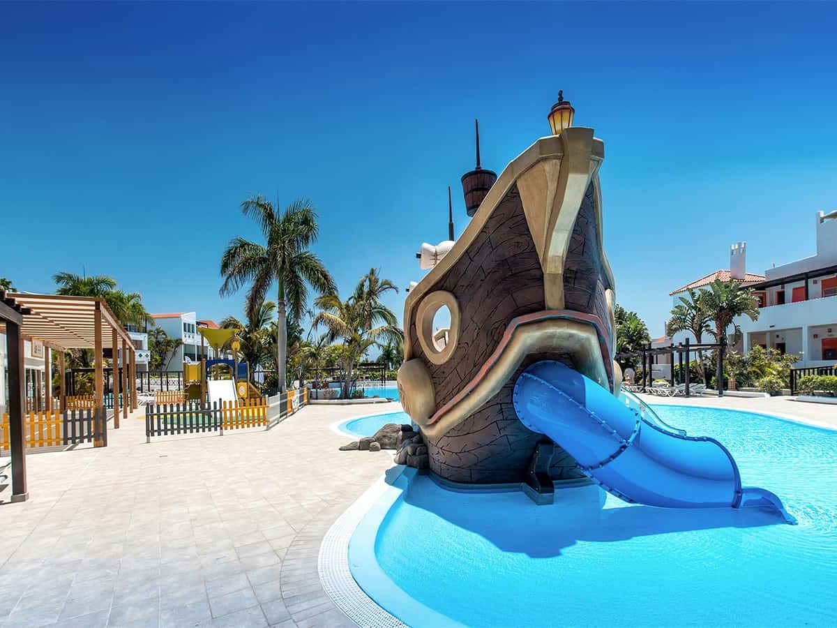 A shallow children's pool with a full-size wooden pirate ship at its centre and a blue water slide running into the water.
