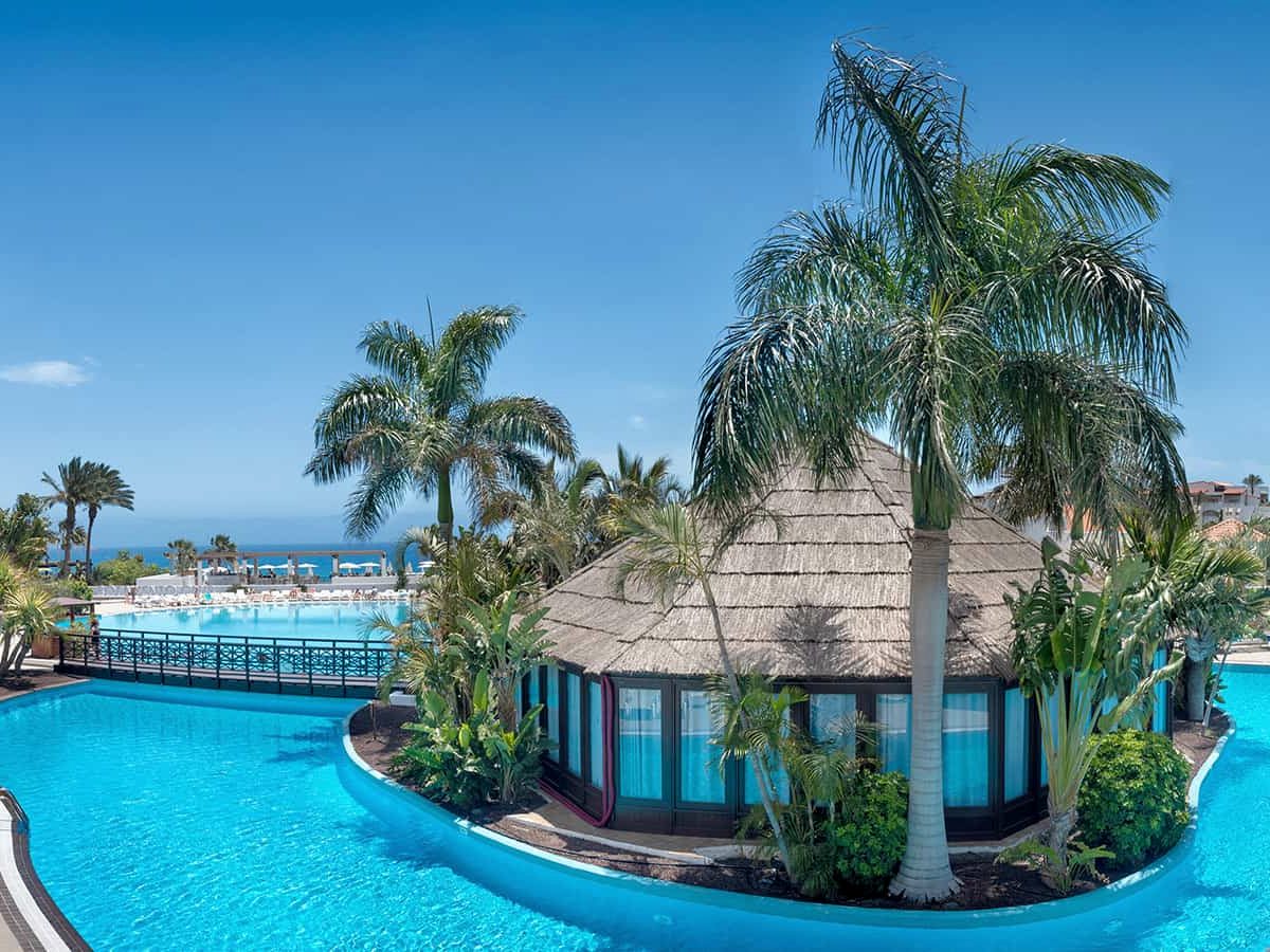 A blue pool curving around a thatched-roof pavilion on a palm-planted island, the sea in the distance.
