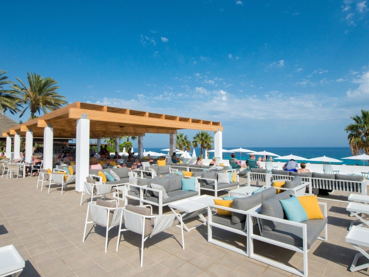 A covered terrace bar with grey lounge sofas and bright cushions under a timber pergola, white parasols and the sea beyond.