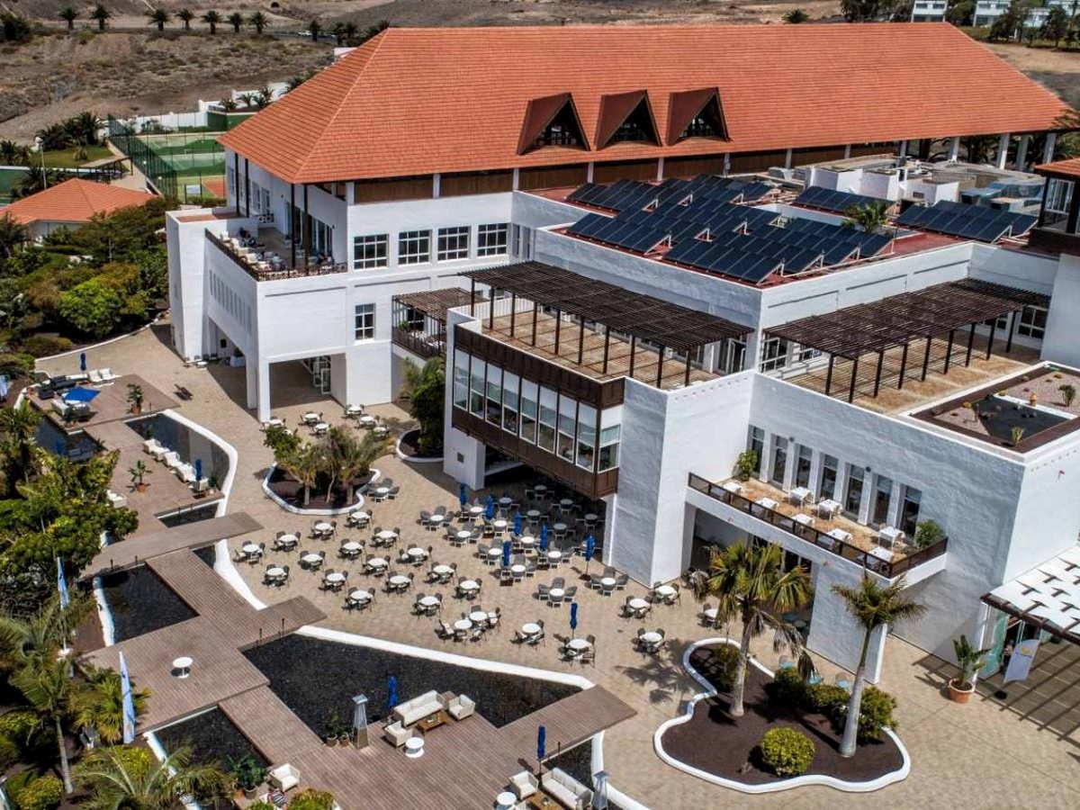 The resort's main building with a terracotta hipped roof, a wide paved terrace of dining tables under blue umbrellas and dark ornamental water channels beside it.