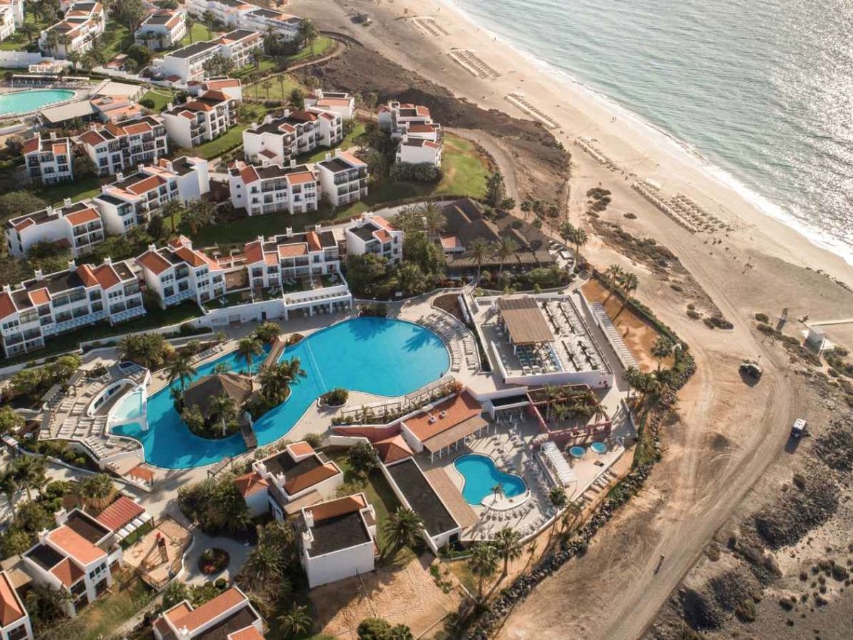 Aerial view of the resort's white buildings and pools beside a long sweep of golden beach and blue sea.