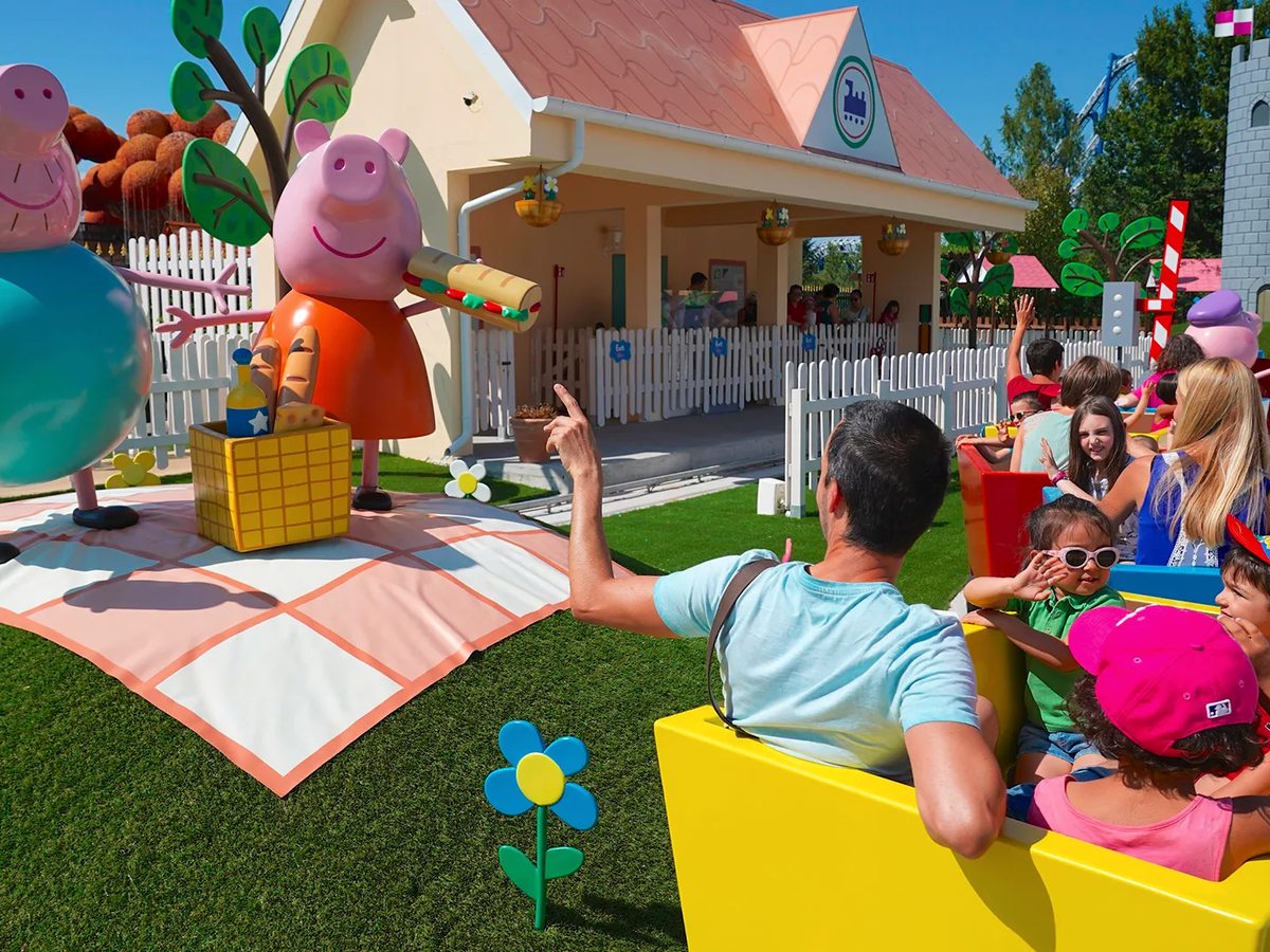 Children riding a car through Peppa Pig Land, with Peppa and Daddy Pig figures and a playhouse facade behind.