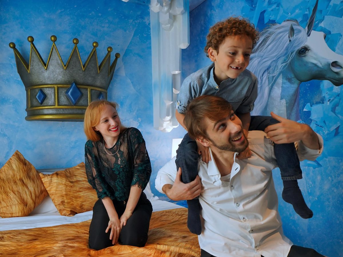 A Magic Ice Castle themed room with a large bed, blue ice-effect walls, a crown wall feature and a unicorn mural, with a family beside it.