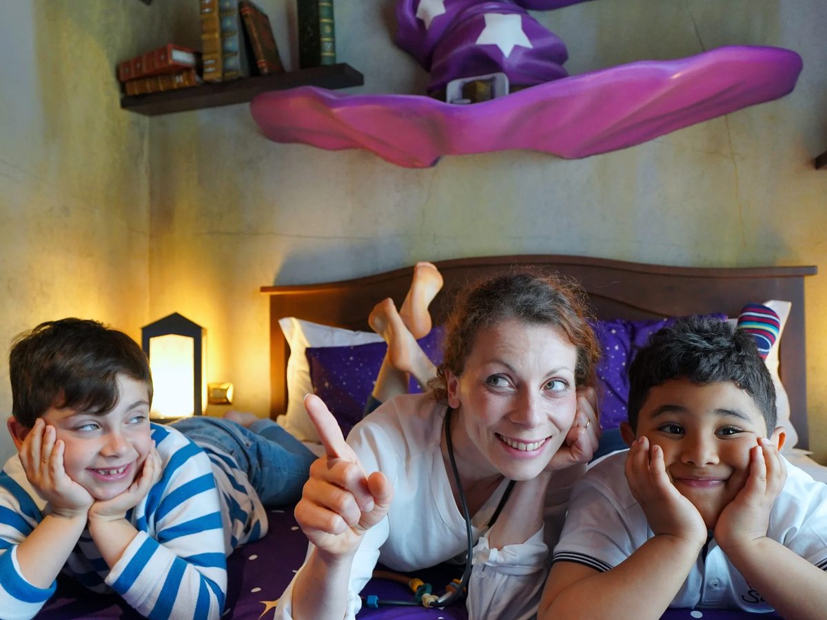 A Wizard House themed room with purple star bedding, a flying wizard's hat wall feature and painted floating books, with children on the bed.