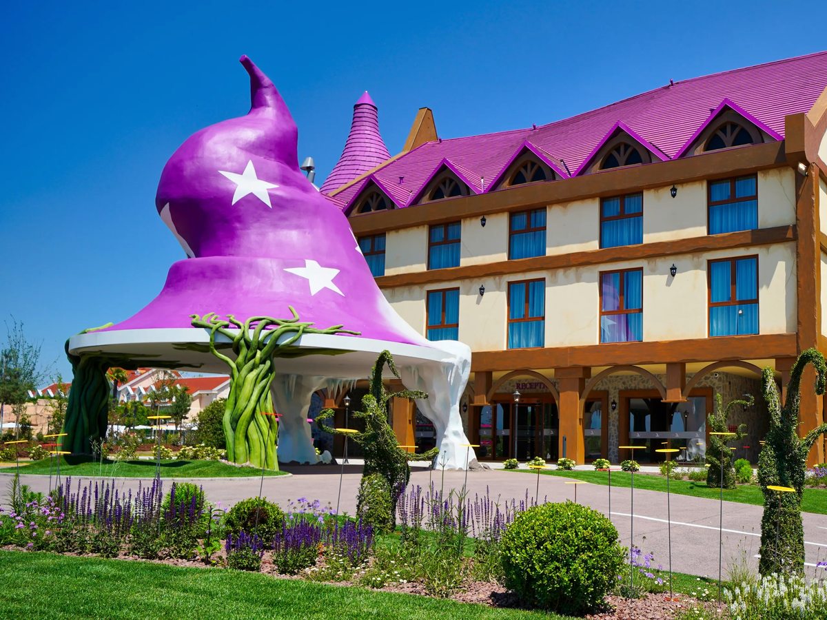 The Gardaland Magic Hotel exterior with a giant purple wizard's hat sculpture over the reception entrance and a magenta pitched roof, above lawns and flowerbeds.