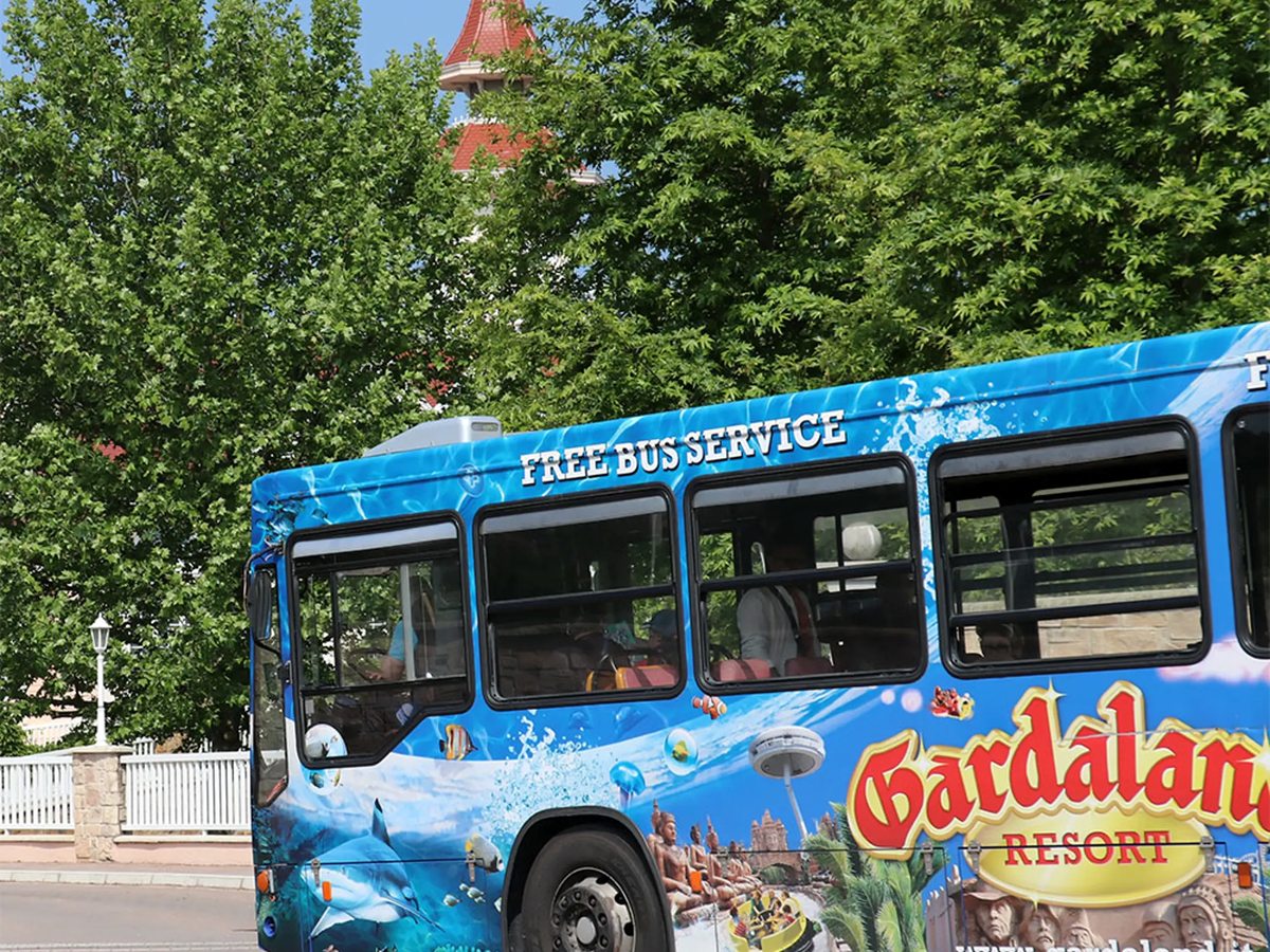 A blue Gardaland Resort shuttle bus marked Free Bus Service, parked at a tree-lined roadside stop.