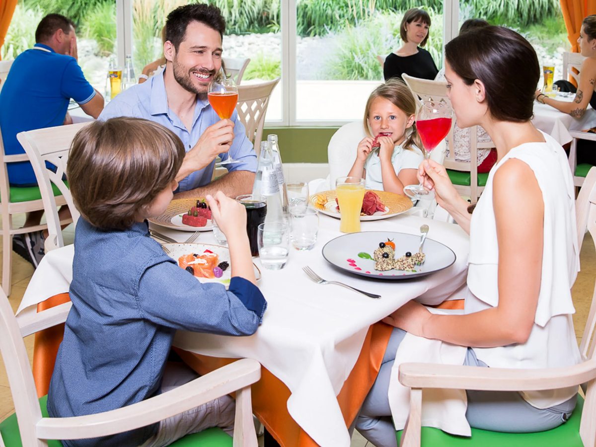 A family at a white-clothed table in a bright, garden-view restaurant with green-cushioned chairs and large windows.