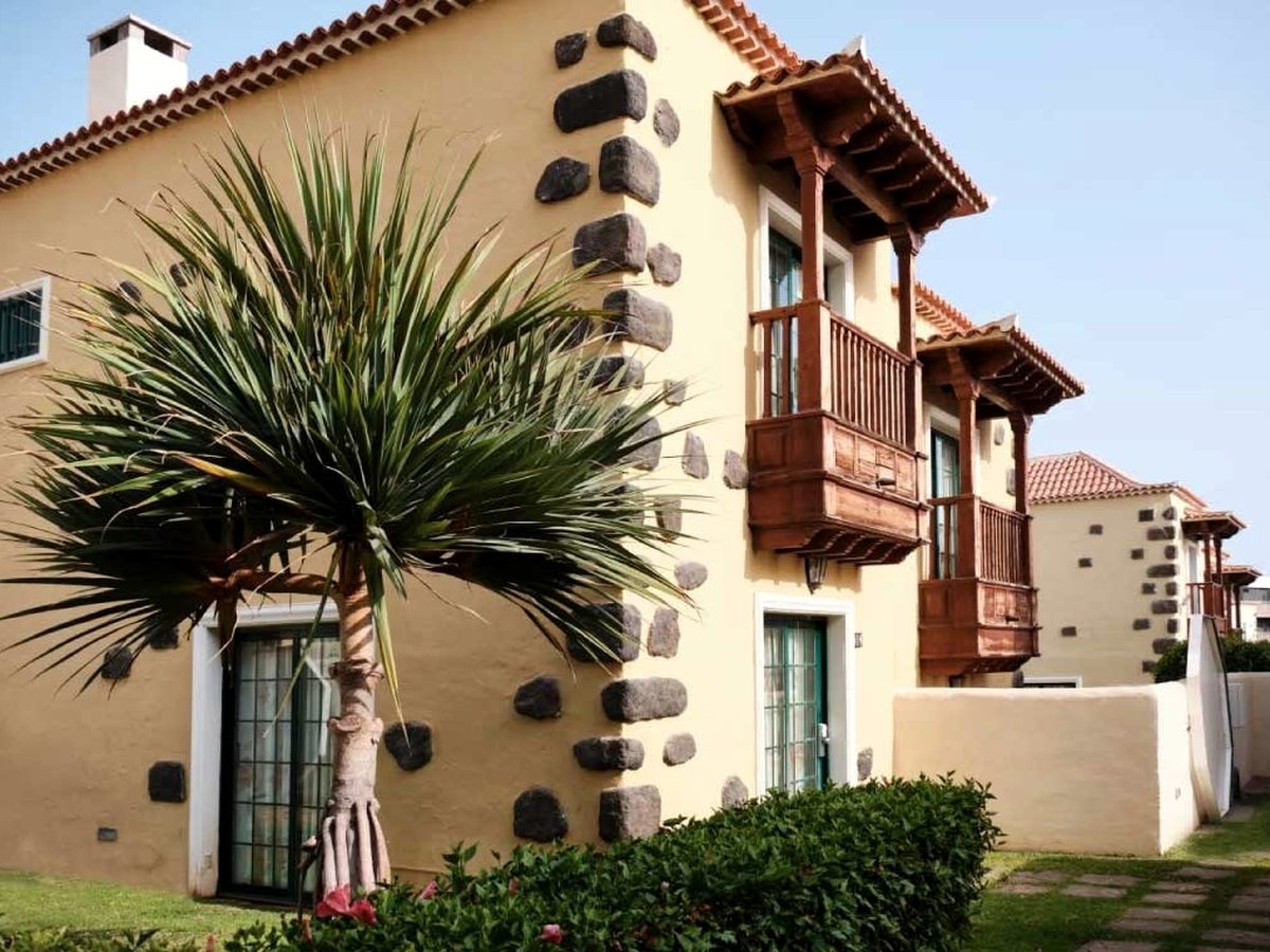 A two-storey villa in cream stucco with volcanic-stone corner detailing, carved wooden Canarian balconies and a terracotta-tiled roof, beside a hedged garden.