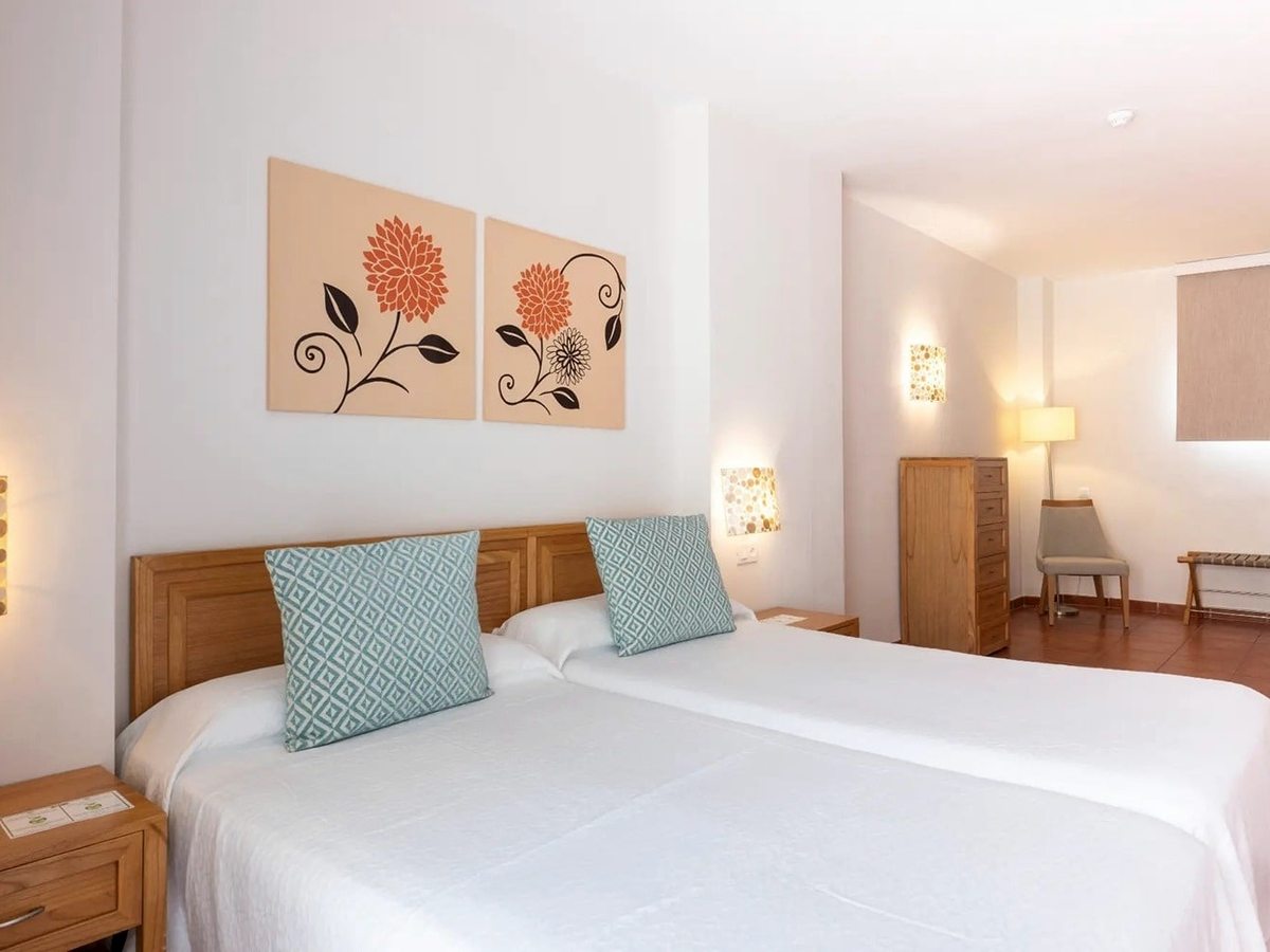 A bright bedroom with two single beds under a wooden headboard, patterned cushions, floral canvas prints, a chest of drawers and an armchair.