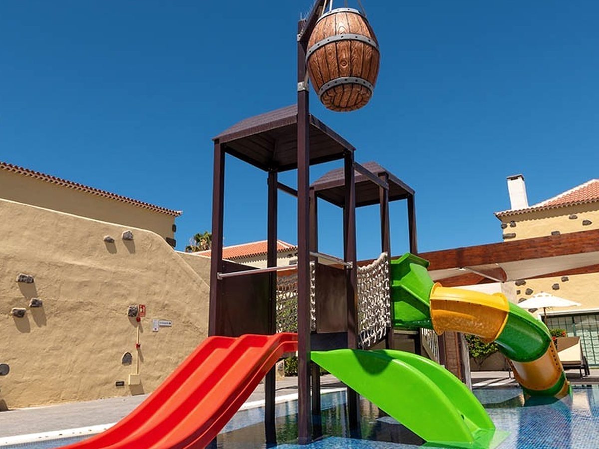 A children's splash pool with a red slide, a green-and-yellow tube slide, a tipping wooden barrel and a climbing wall, against cream village buildings.