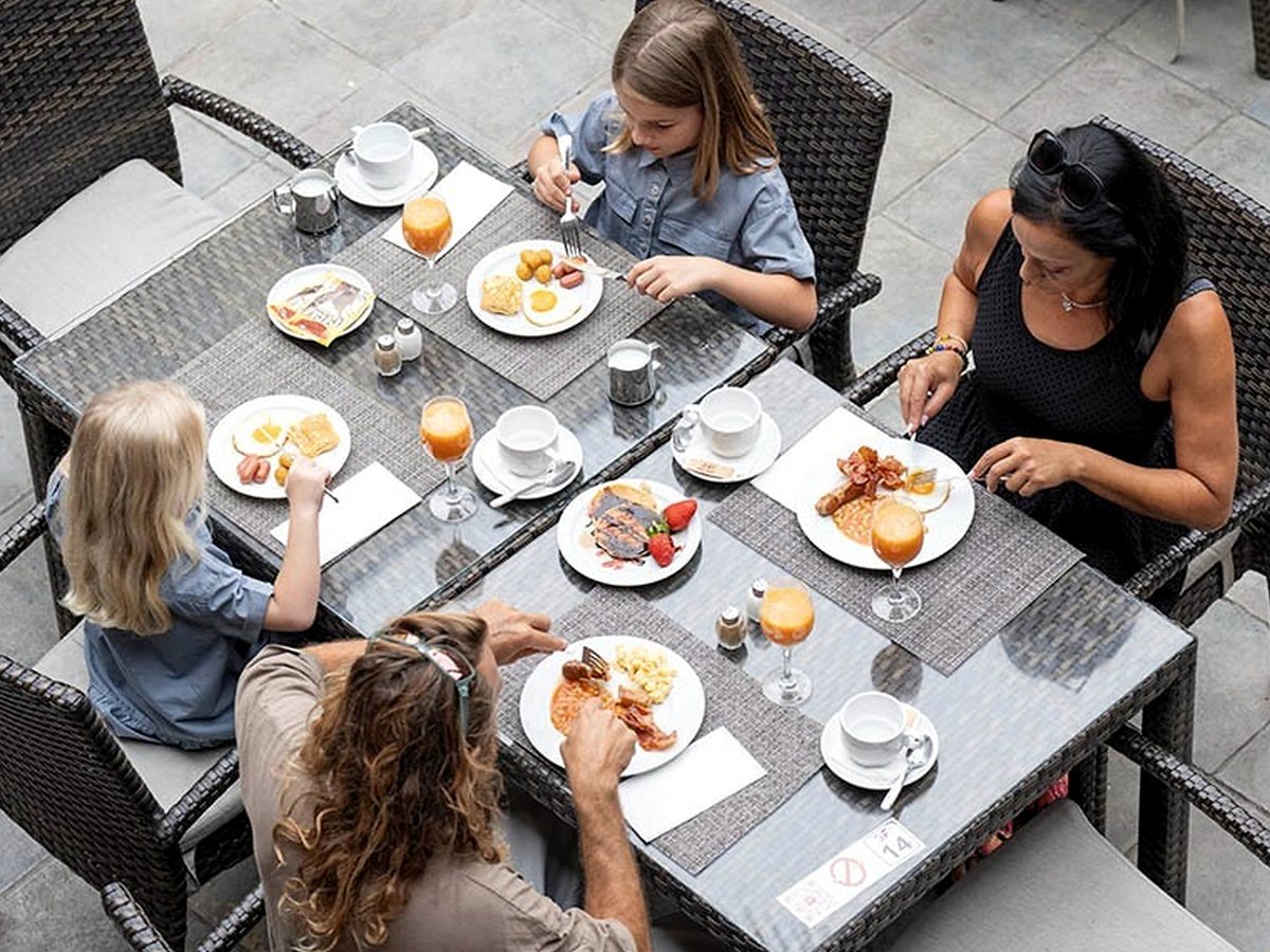 A family of four at a table on the paved terrace, with plates of cooked breakfast, pastries and glasses of orange juice, seen from above.