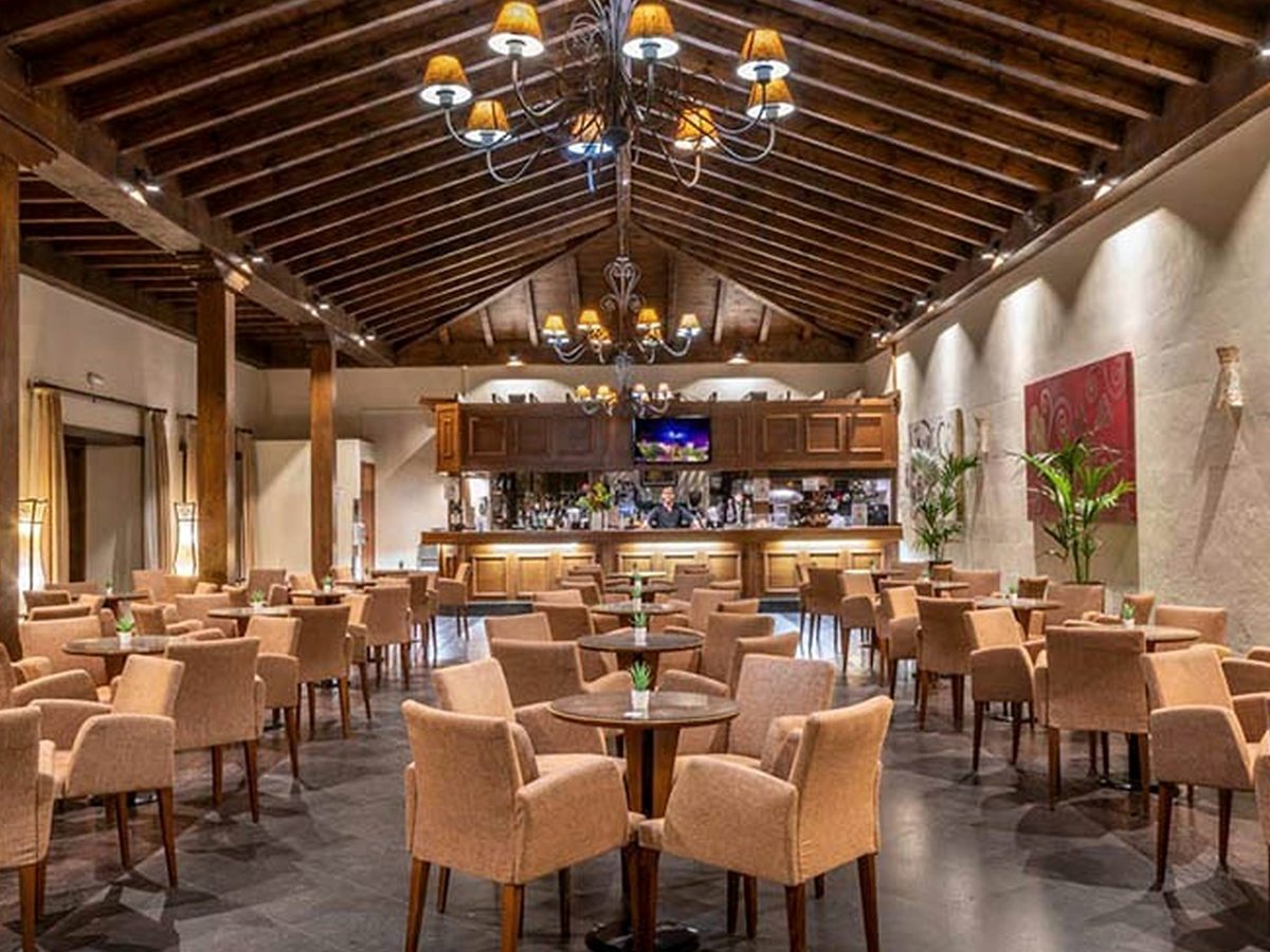 A rustic dining room with a high timber-beamed ceiling, wrought-iron chandeliers, upholstered armchairs at round tables and a wooden bar counter at the far end.