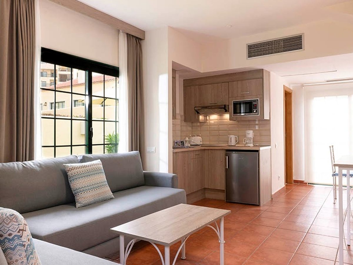 An open-plan living area with a grey sofa and a corner kitchenette with wood units, a microwave, a fridge, a kettle and a coffee maker, on a terracotta-tiled floor.