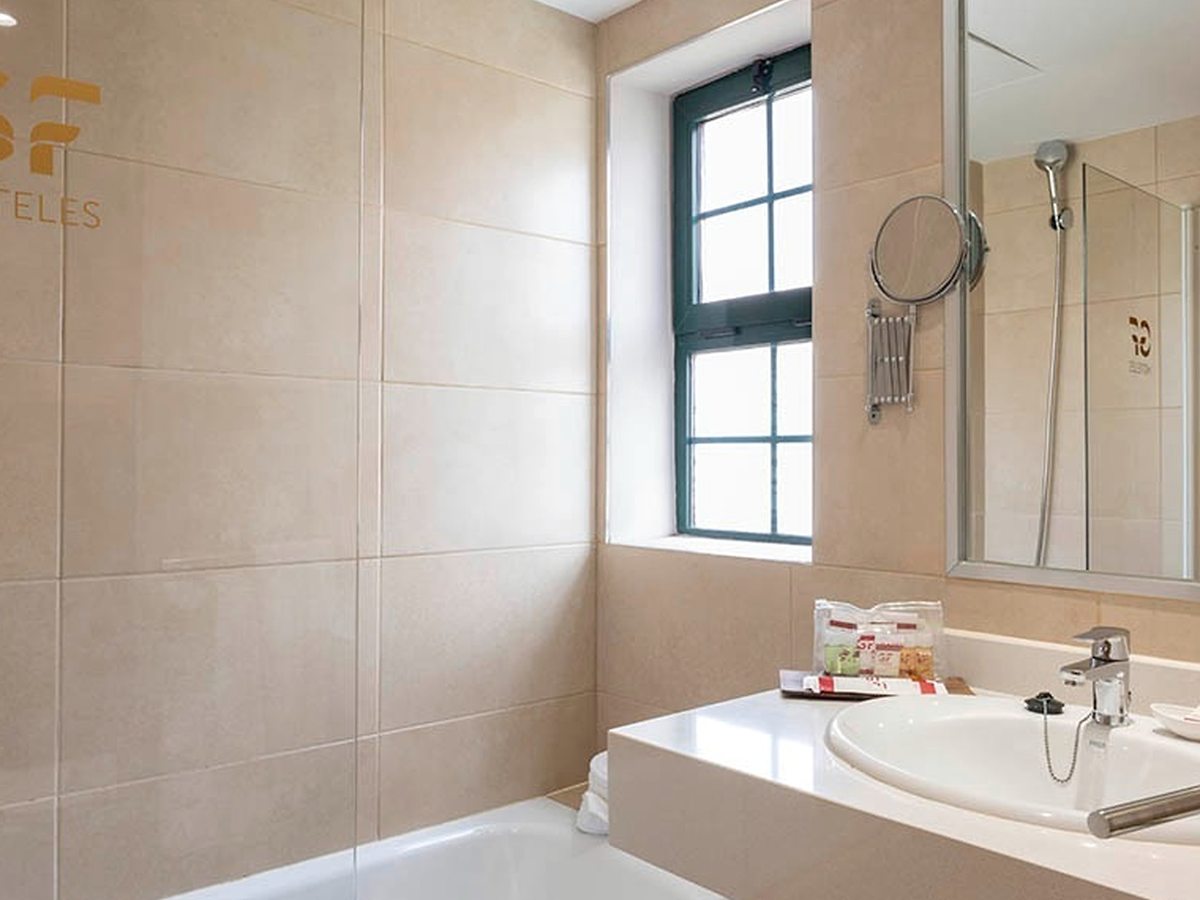 A beige-tiled bathroom with a bathtub below a green-framed window, a vanity unit with a basin, a large mirror and a wall-mounted hairdryer.