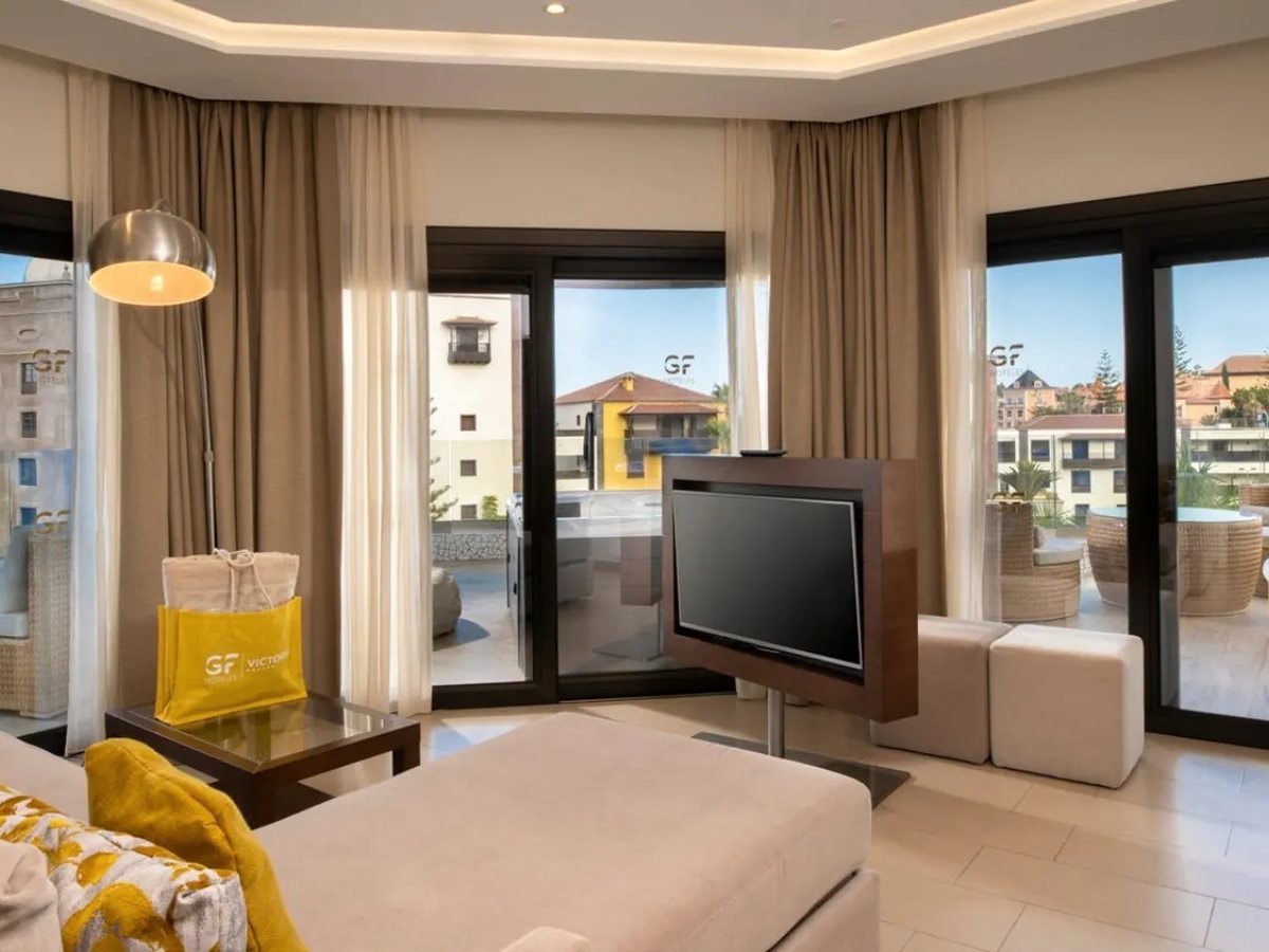 A bright suite lounge with a cream sofa, arc floor lamp and swivel TV, sliding doors onto a furnished terrace.