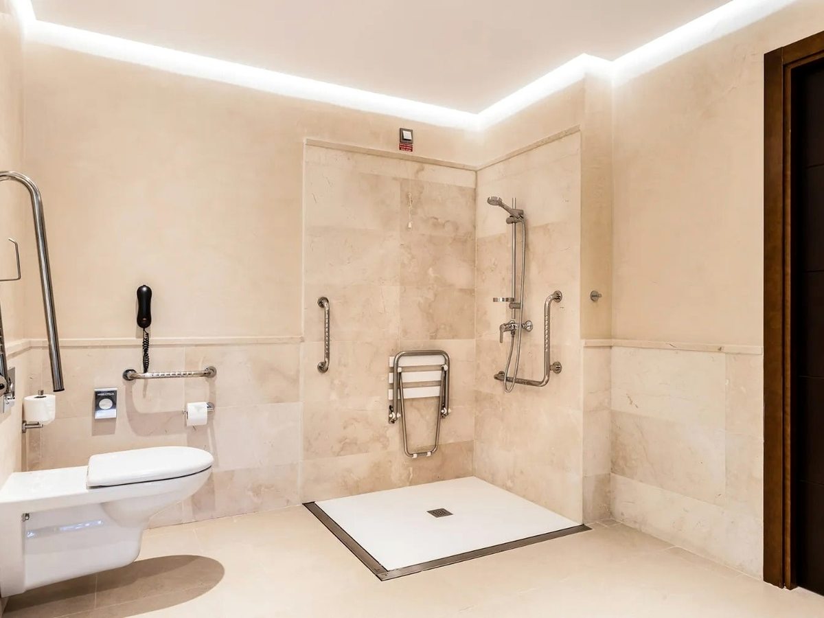 A marble wet-room bathroom with a level-entry shower, fold-down seat and grab rails beside a wall-hung toilet.