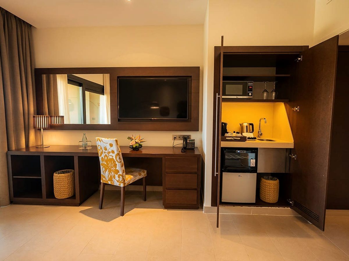 An open dark-wood cabinet holding a microwave, kettle, mini-fridge and Nespresso machine beside the desk.