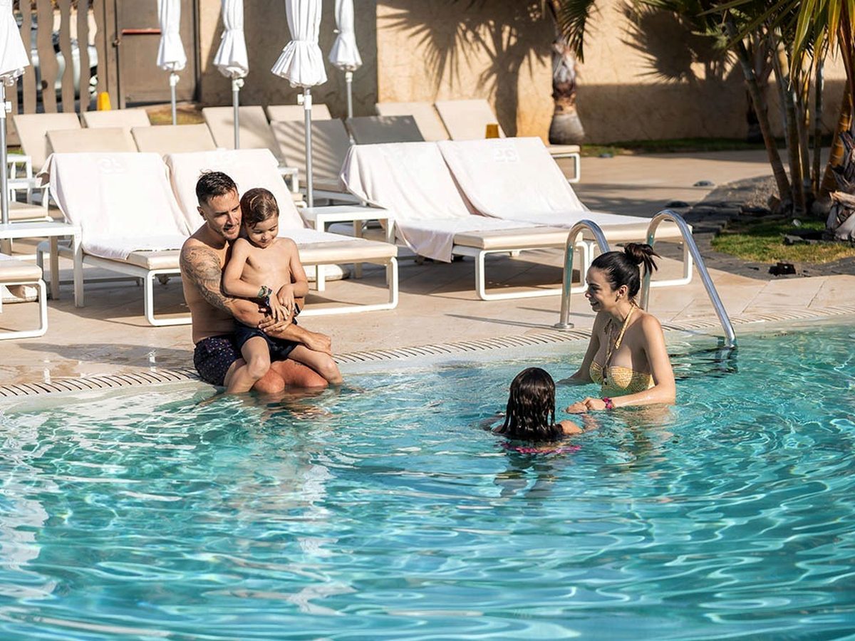 Two parents and two children playing in a shallow blue pool edged with white loungers and palms.