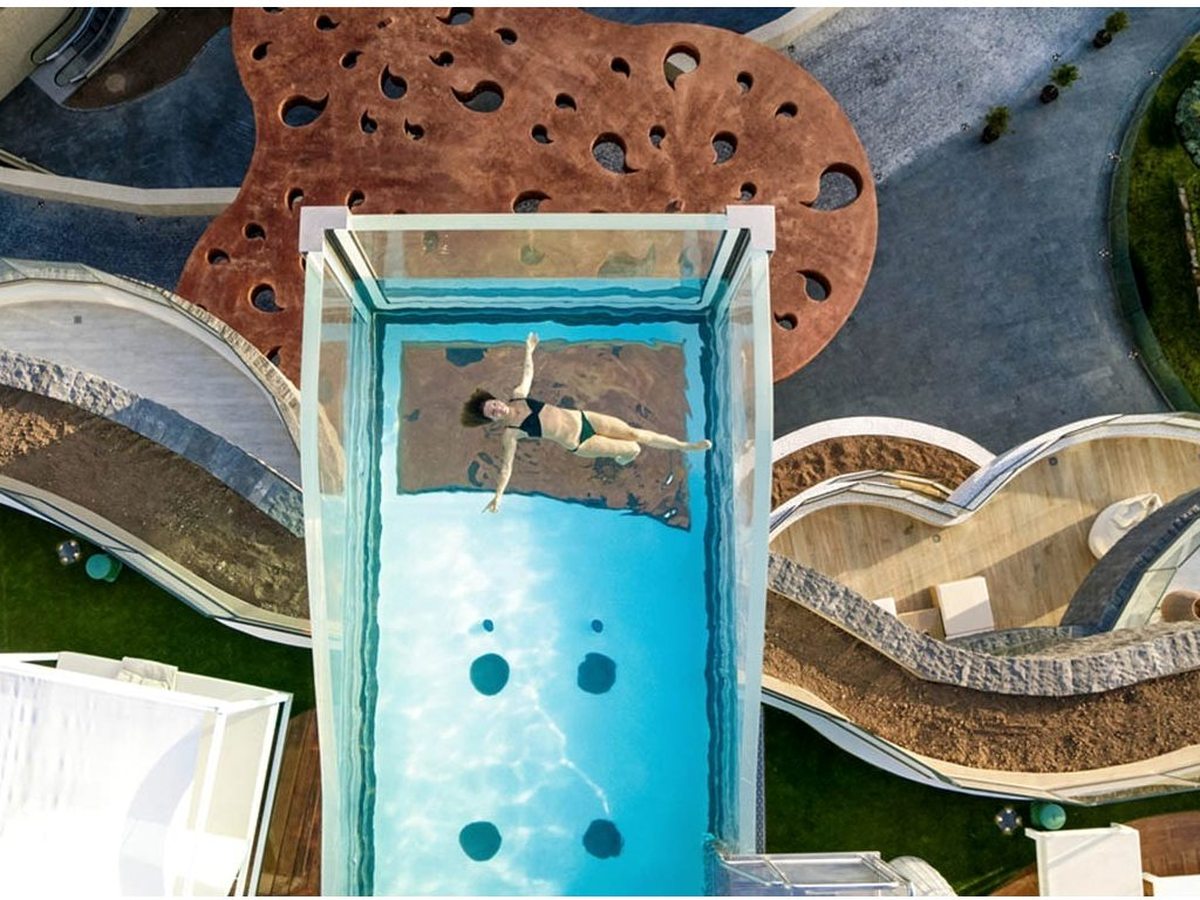 An overhead view of a glass-walled pool projecting from the building, a swimmer floating on the surface.
