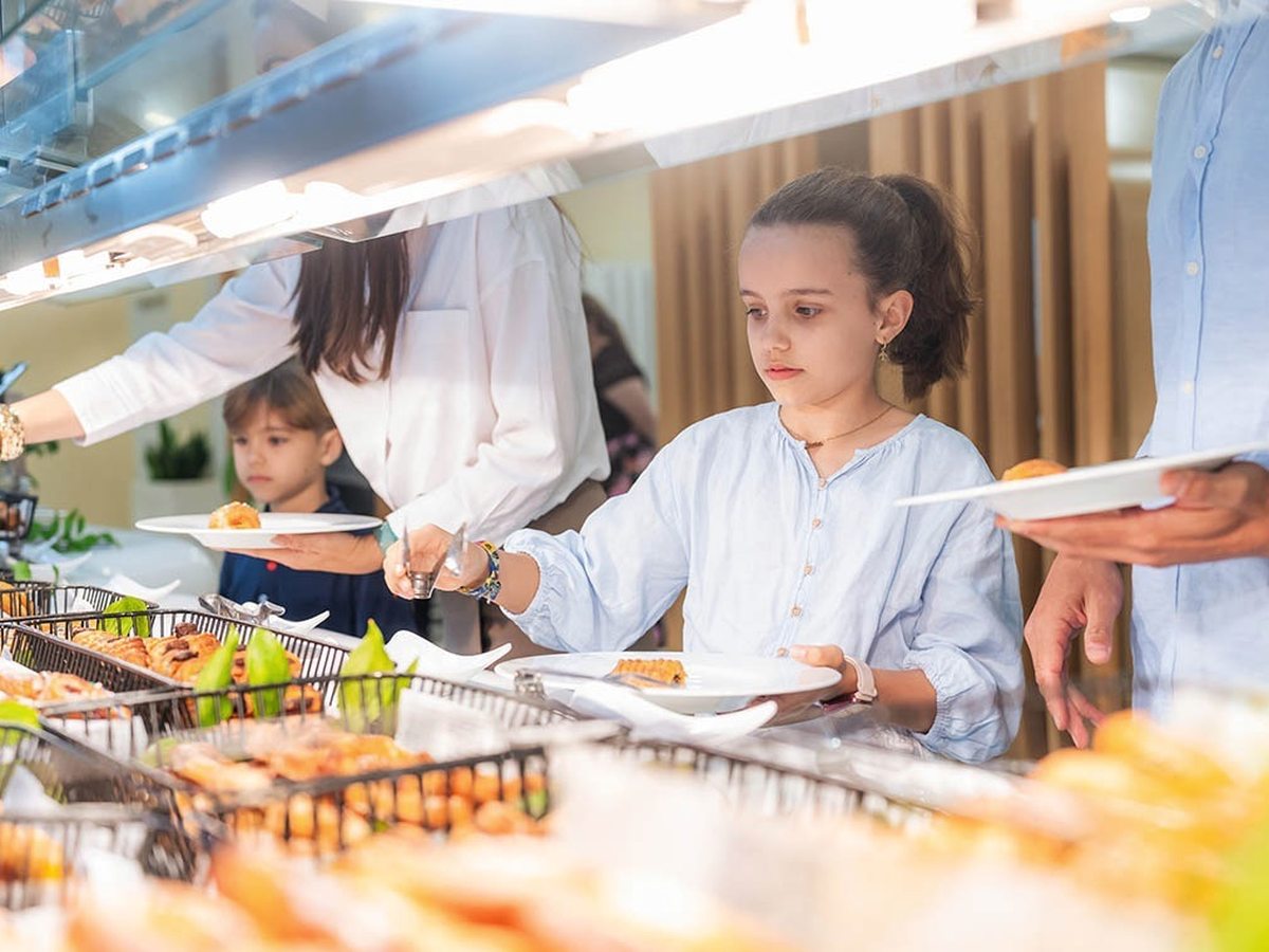 Two children choosing pastries from a bright buffet, each holding a white plate, a parent alongside.