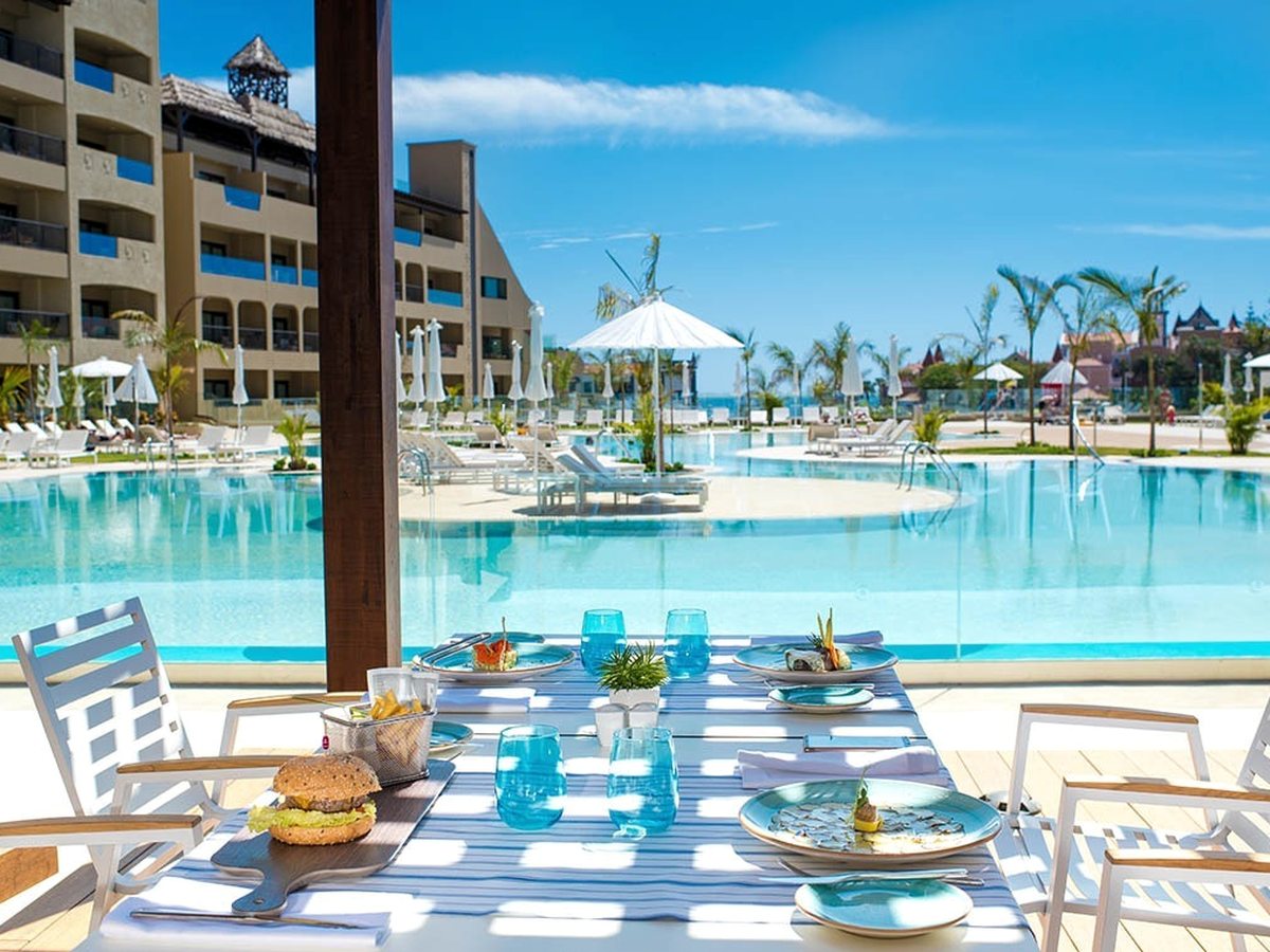 A poolside table set with plated food and blue glasses, the resort pool and a hotel wing beyond.