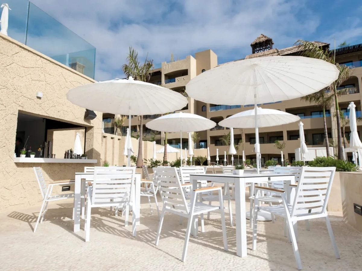 White parasols, tables and chairs on a paved sun terrace with the tan hotel building and palms behind.