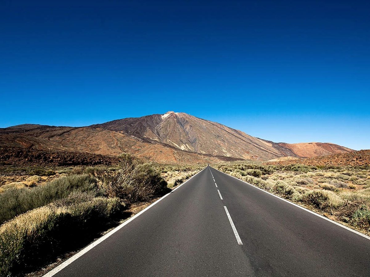 The snow-flecked summit of Mount Teide at the end of a straight empty road through volcanic scrubland.