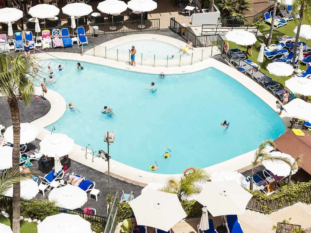 An aerial view of a large curved family pool with swimmers, an attached shallow section, blue loungers and white parasols around it.