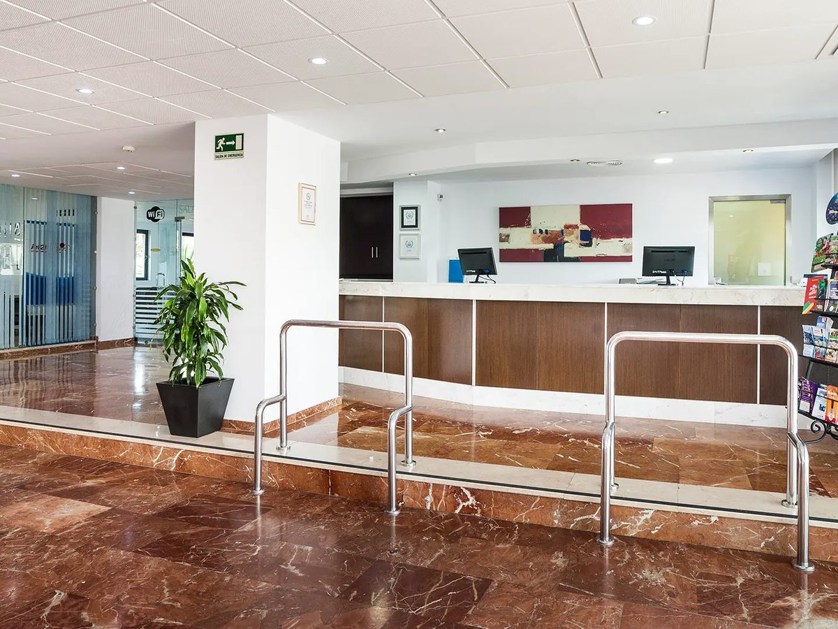 The hotel reception desk with a wooden front and a ramp with metal railings running up to it across a marble floor.