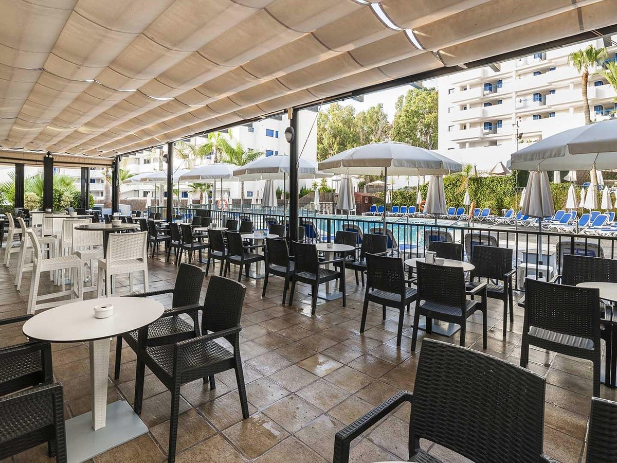 A covered poolside terrace with black wicker chairs and round tables, looking out over the pool to the white hotel block.