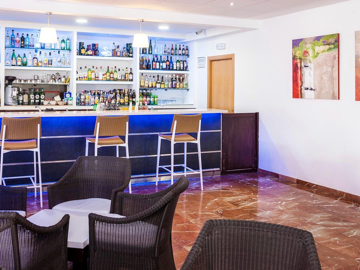 An indoor bar with a blue-lit counter, shelves of bottles behind, bar stools and low wicker armchairs on a marble floor.