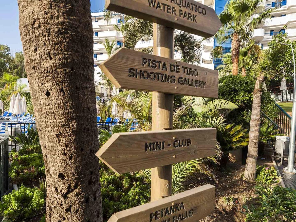 A wooden signpost fixed to a palm trunk, with painted arrows for the water park, shooting gallery, mini club and petanque.