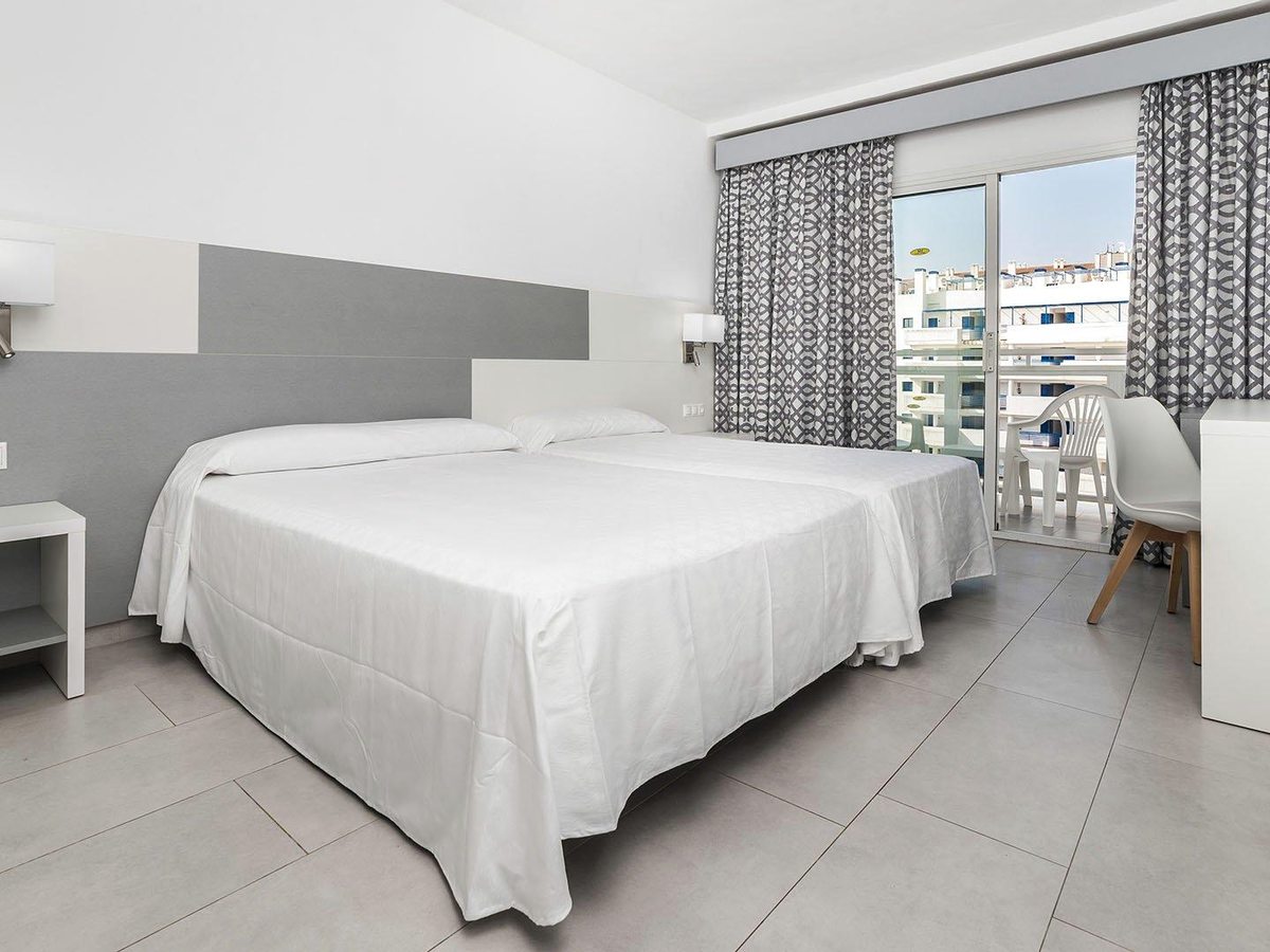 A double room with a balcony behind patterned curtains, white bedding, a grey headboard and a tiled floor.