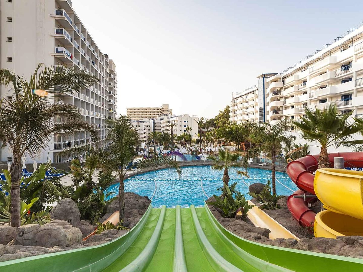 An elevated view looking down green water slides towards a large round pool, with red and yellow tube slides to one side and the white hotel blocks rising on either side.