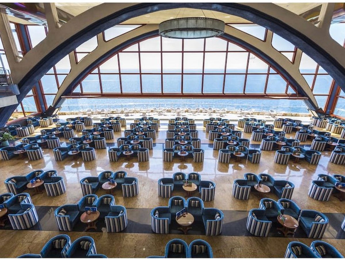 Large buffet restaurant hall with rows of blue tub-chair booths under a tall arched glass wall looking out to the ocean.