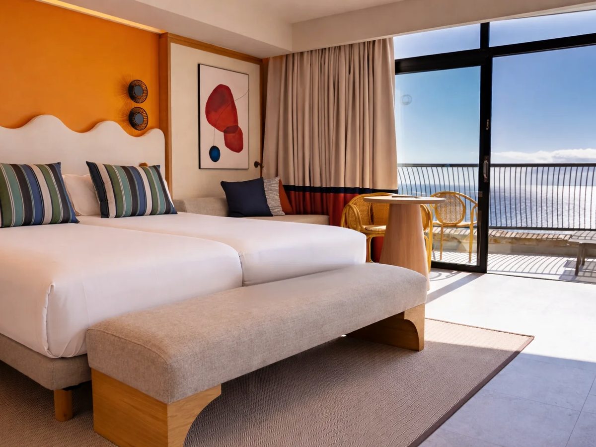 Hotel double room with an orange headboard wall, two beds pushed together, and a glazed balcony looking out over the sea.
